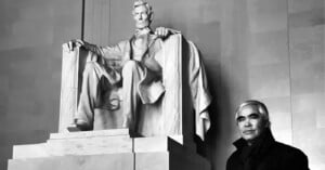 A person stands beside the large marble statue of Abraham Lincoln at the Lincoln Memorial. The statue shows Lincoln seated in a chair, looking serious. The person wears a coat and scarf, and both the statue and the person are in a dimly lit setting.