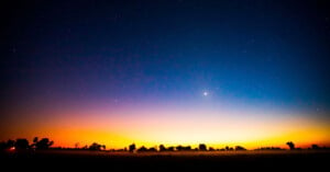 A vibrant twilight sky with a gradient from deep blue to orange at the horizon, scattered with stars. A bright celestial object is visible slightly above the horizon. Silhouettes of trees line the landscape.