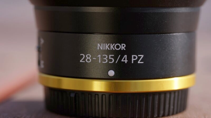 Close-up of a camera lens showing the text "NIKKOR 28-135/4 PZ" in white, with a gold ring around the base of the lens. The focus is on the text and the black and gold design.