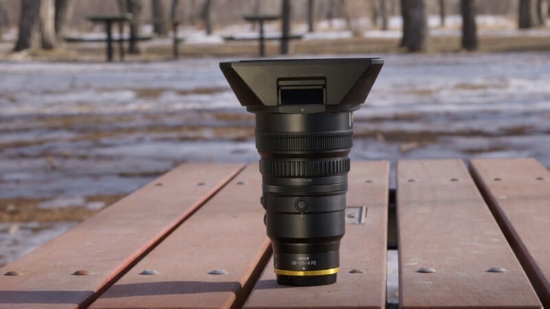 A camera lens with a wide hood stands upright on a wooden picnic table. The background features a park setting with leafless trees and snow patches on the ground. The lens labels indicate it is a 28-70mm f/2 model.