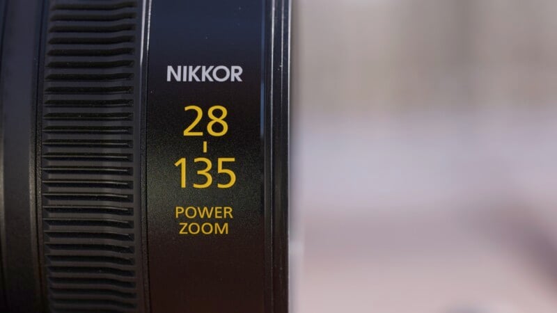 Close-up of a Nikkor camera lens, showing the text "28-135 POWER ZOOM" in yellow on a black background, indicating the focal length range and zoom capability.