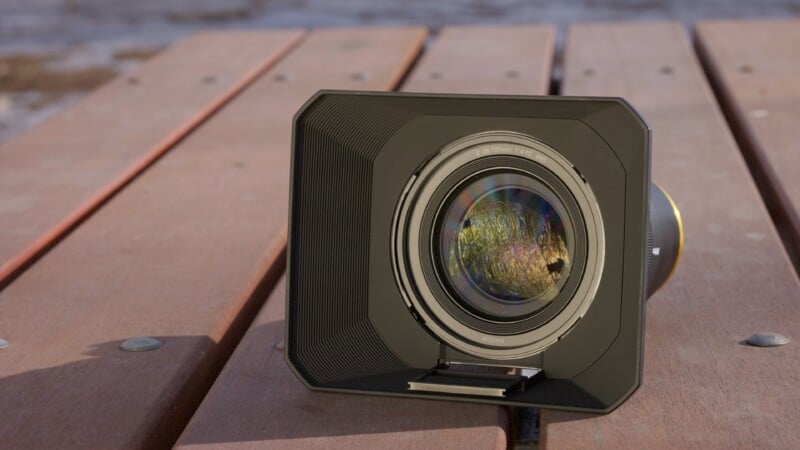 A camera lens with a rectangular lens hood sits on a wooden surface. The lens reflects a colorful, abstract pattern, possibly a sunset or landscape. The background is blurred, suggesting an outdoor setting.