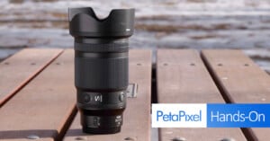 A camera lens with a hood is standing upright on a wooden surface outdoors. The background is blurry, showing hints of water. The lens has various markings and logos. The bottom right corner displays "PetaPixel Hands-On" in a white and blue banner.