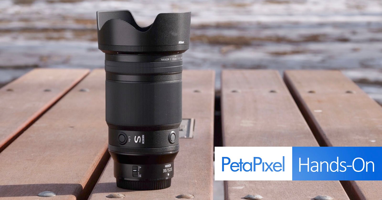 A camera lens with a hood is standing upright on a wooden surface outdoors. The background is blurry, showing hints of water. The lens has various markings and logos. The bottom right corner displays "PetaPixel Hands-On" in a white and blue banner.