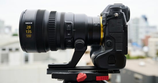 Side view of a professional camera with a large zoom lens mounted on a tripod. The lens has "28 135" and "Power Zoom" marked on it. The background is a blurred urban landscape, suggesting an outdoor setting.