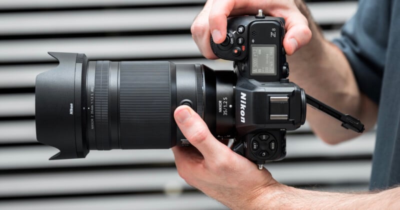 Close-up of a person holding a Nikon camera with both hands. The camera has a large lens and a digital display screen showing settings. The background features horizontal grey stripes.