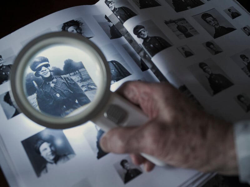 A hand holds a magnifying glass over a black-and-white photograph in a book filled with historical images. The photo under the magnifying glass shows a person in a military uniform. The surrounding images are similar in style and theme.