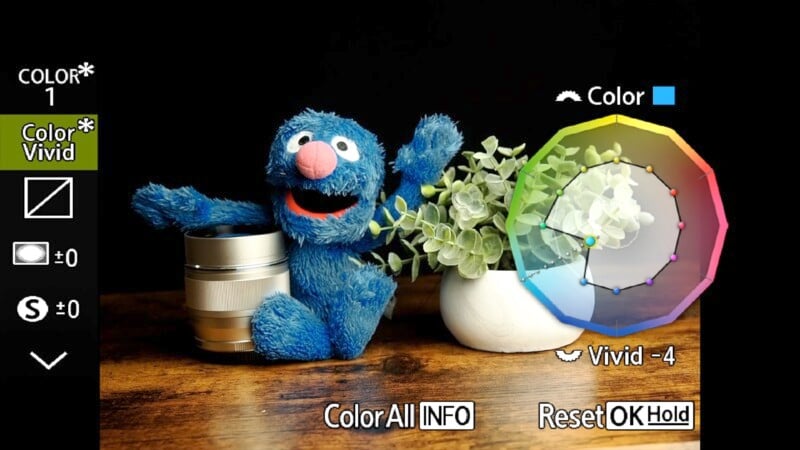 A blue, furry puppet next to a camera lens sits on a wooden surface. To the right, a white vase holds green leafy plants. The digital display shows color adjustment options on the screen.