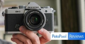 Person holding a vintage silver and black OM-3 camera with a M.Zuiko Digital lens. The image features a "PetaPixel Reviews" graphic in the bottom right corner. Background is blurred.