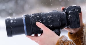 Close-up of a person holding a black camera with a large lens, covered in snowflakes. The background is snowy and blurred, highlighting the winter setting. The person is wearing a brown jacket.