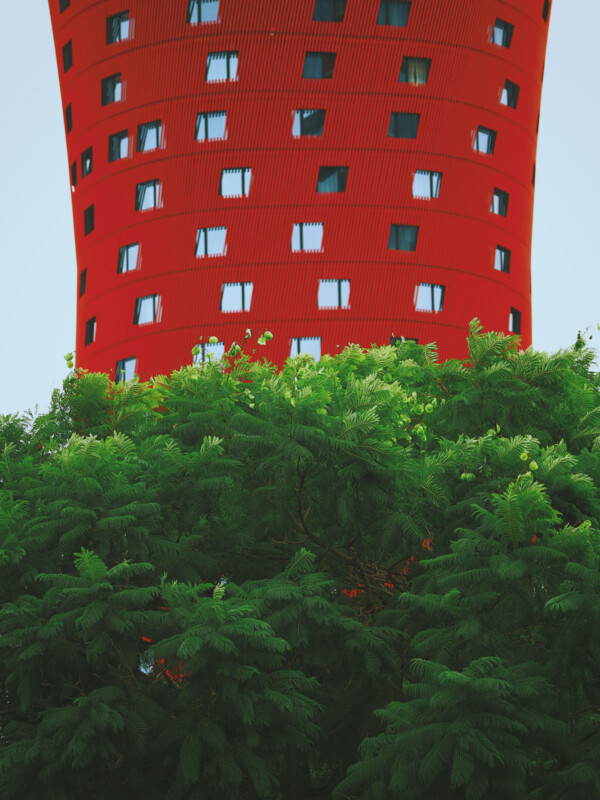 A unique building with a red, cylindrical shape featuring square windows looms above lush, green foliage. The structure creates a striking contrast between the vibrant red facade and the vivid greenery below.