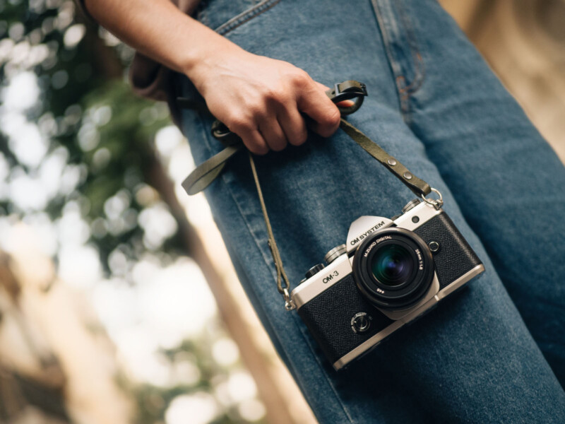 Close-up of a person holding a vintage-style camera by its strap. The person is wearing blue jeans, and the camera, which is black and silver, hangs by their side. The background is slightly blurred, with hints of greenery and a building.