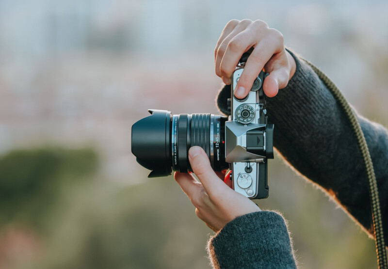 Close-up of a person holding a vintage-style camera with both hands. The individual is wearing a dark sweater. The camera features a silver body and a large lens. The background is softly blurred, showcasing natural greenery.