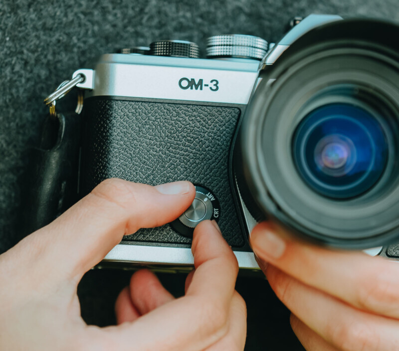 Close-up of hands adjusting the settings on a vintage camera labeled "OM-3." The camera has a textured, black grip and a large lens, with fingers turning a dial on the top. A woolen surface is visible in the background.