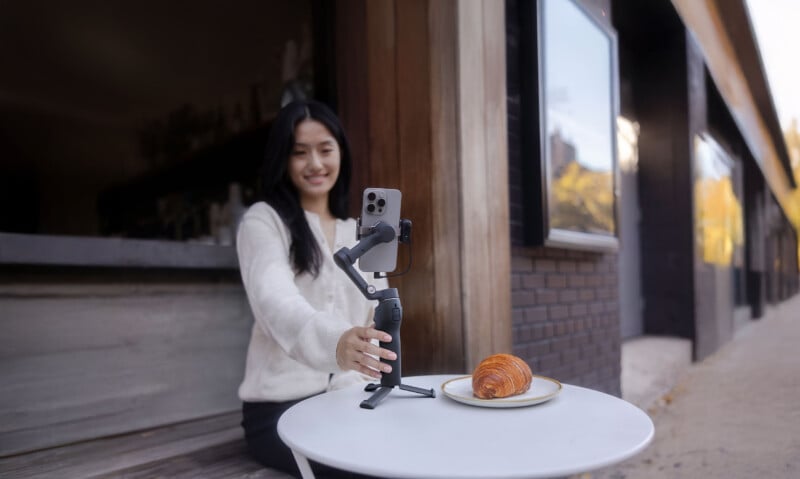 A person is sitting at an outdoor table, smiling at a smartphone mounted on a stand. A croissant is on a plate in front of them. The background features a building with large windows.