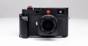 A black Leica camera with a textured grip and a prominent lens is placed on a white surface. It features a red Leica logo and various dials and buttons on top. The design is sleek and professional.