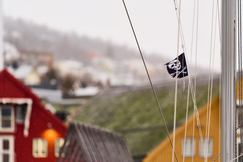 A black pirate flag with a skull and crossbones design is hoisted on a mast. In the blurred background, colorful buildings including red and yellow structures are visible, suggesting a coastal or harbor setting on a cloudy day.