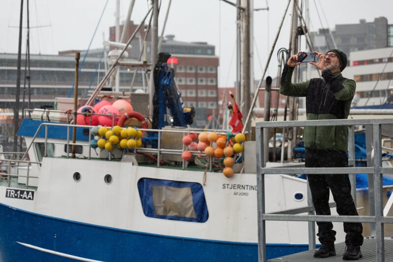 A person in warm clothing stands on a dock, taking a photo with a smartphone. Behind them is a fishing boat adorned with colorful buoys. The background features city buildings under an overcast sky.