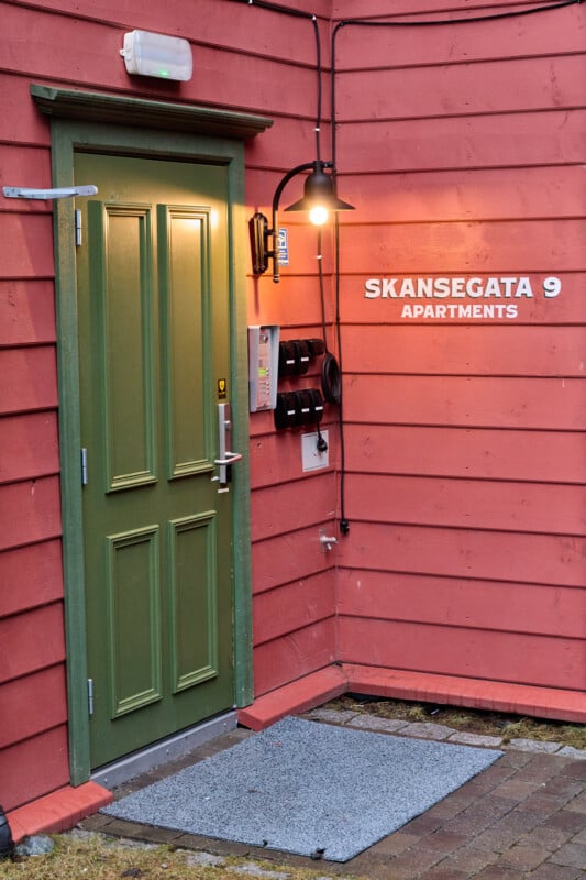 A green door is set in a salmon-colored wall, illuminated by a vintage outdoor lamp. The wall displays the text "Sk​​ansegata 9 Apartments." A set of mailboxes and a doorbell panel are mounted beside the door.