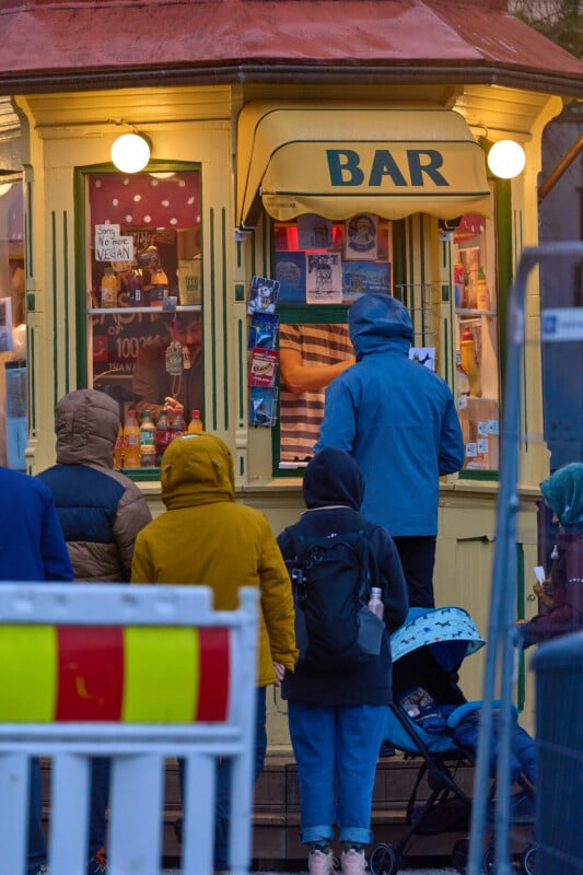 People in hooded jackets stand outside a small yellow kiosk with an awning labeled "BAR." Inside, shelves hold various items. The scene is lively, with signs on the window, and a stroller is visible among the group.