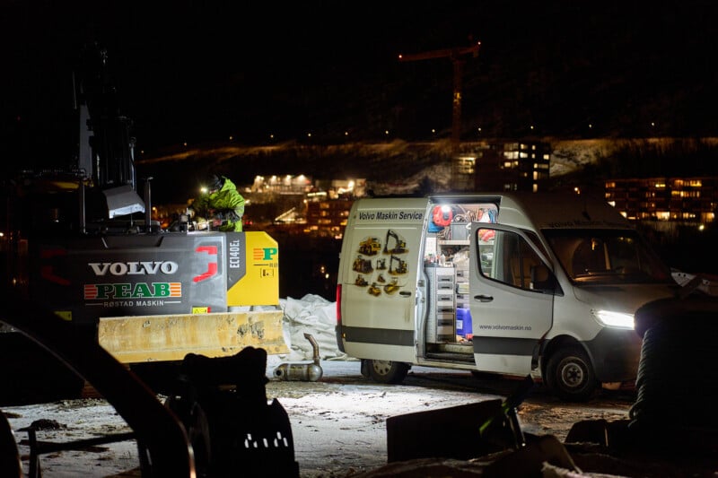 Nighttime construction site with a lit service van parked beside a Volvo excavator. The van's open side displays tools and equipment inside. Snow-covered ground and illuminated buildings can be seen in the background.