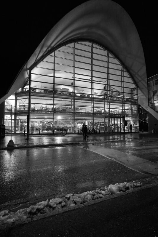 A black and white image of a modern building with a large, curved roof and glass facade. The interior is brightly lit, revealing multiple floors with people, furniture, and vehicles visible through the windows. Wet pavement reflects the structure.