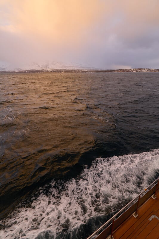 View from a boat showing dark, choppy waters with white foam trailing behind. Snow-covered mountains and a distant shoreline are visible under a cloudy, orange-tinted sky during sunset or sunrise.