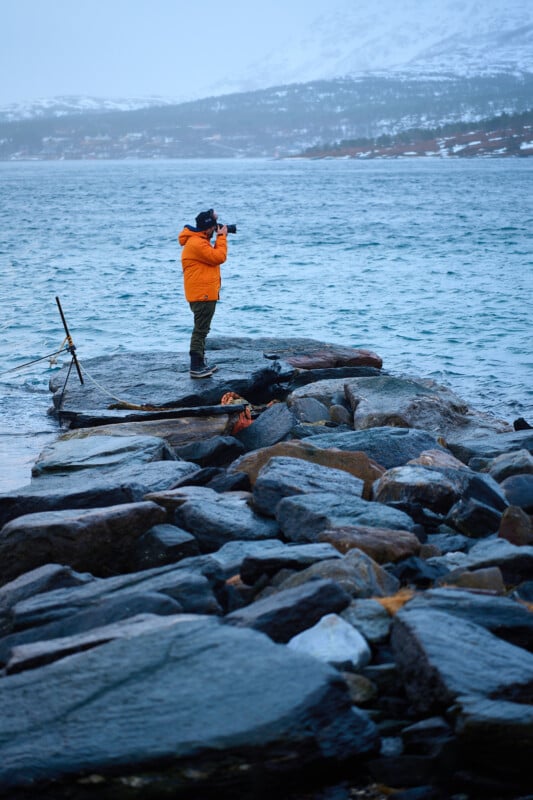 A person in an orange jacket stands on a rocky shoreline taking a photo of the sea with mountains in the background. The water is calm, and the sky is overcast.