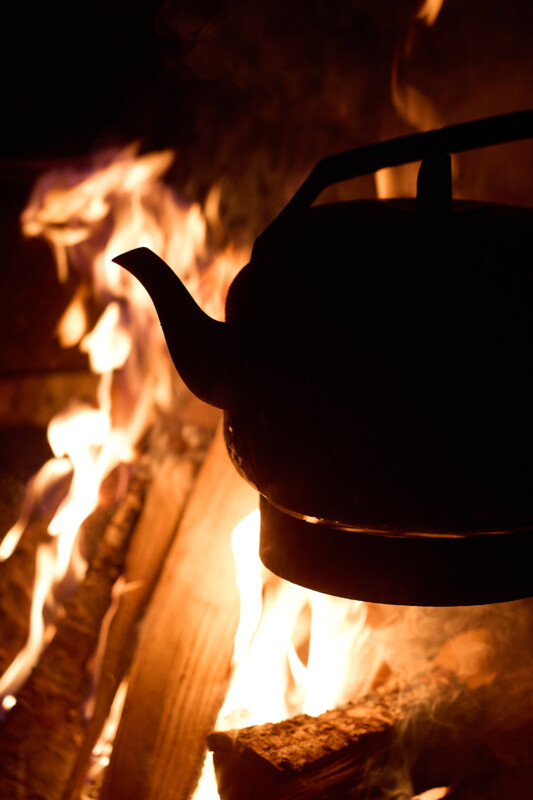 A silhouette of a kettle is seen suspended over a roaring fire. The flames are bright and flickering, illuminating the kettle's outline against the dark background. Logs underneath the fire are partially visible.