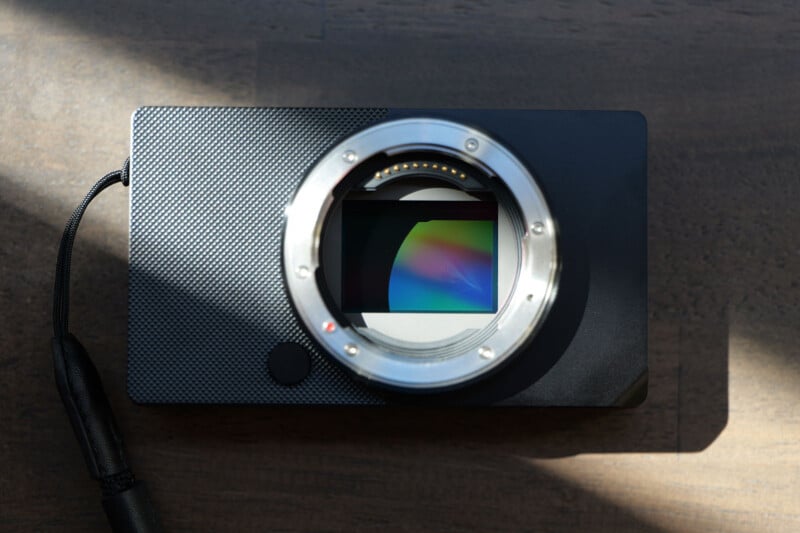 A compact digital camera body with no lens attached is placed on a wooden surface. The camera's sensor is visible through the lens mount. Sunlight casts shadows across part of the camera.