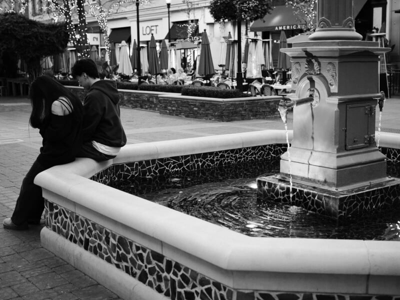 A couple sits back-to-back on the edge of a decorative fountain in an outdoor plaza. The scene is in black and white, with string lights and store signs visible in the background.