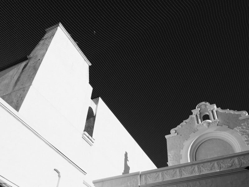 Black and white photo of architectural details with sharp angles and curves, highlighting a bell tower and an ornate façade under a netted sky. The contrast creates a striking visual with deep shadows and bright surfaces.