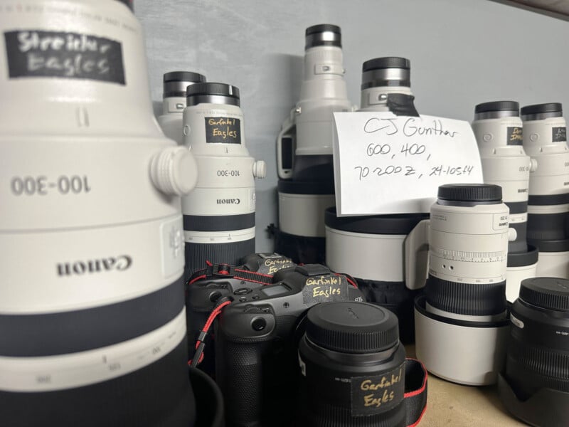 A collection of professional Canon camera lenses and camera bodies are arranged on a surface. Some lenses are labeled "Stacking Eagles." A note on one lens reads, "CJ Gunther, 600, 400, 70-200 2.8, XT-105-4.