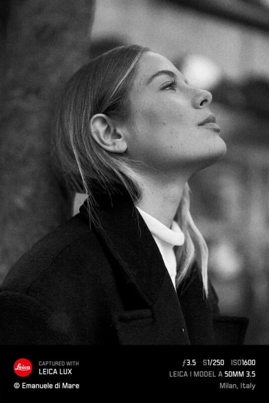 Black and white portrait of a person with long hair, wearing a dark coat and white turtleneck, looking upwards with eyes closed. The image has a soft focus and includes camera settings and location text at the bottom.