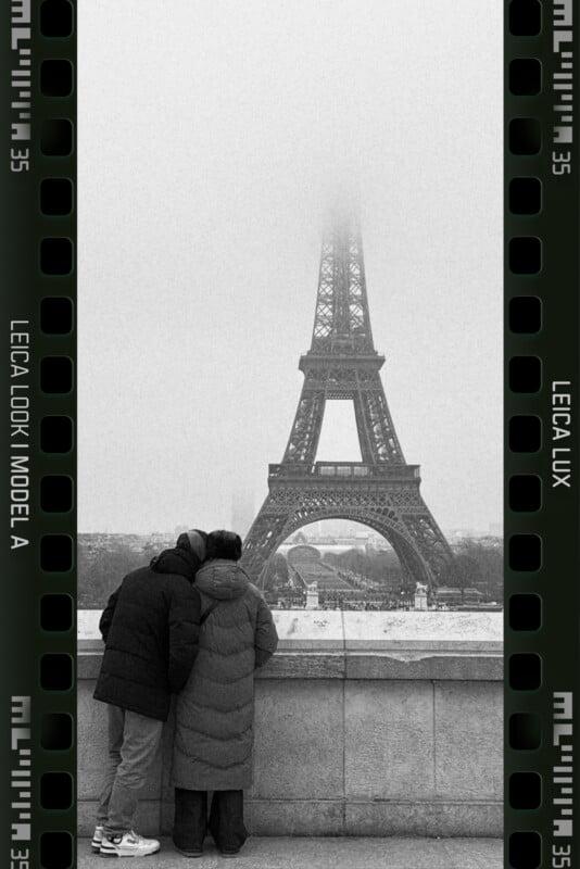 Two people in winter coats lean against a stone railing, looking at the Eiffel Tower obscured by fog. The image is framed with a film strip border, adding a vintage feel.