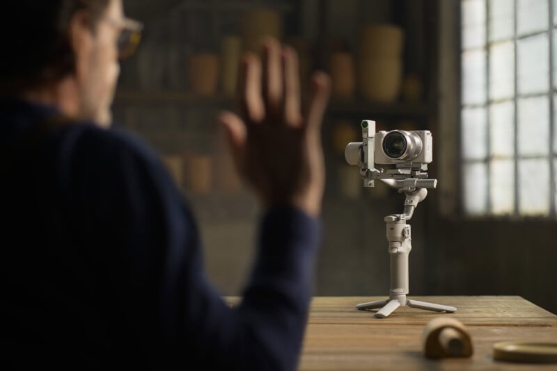 A person gestures in front of a white digital camera mounted on a small tripod with a gimbal, placed on a wooden table. The setting appears to be indoors, with soft lighting and shelves in the background.