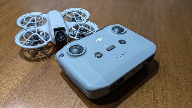 A compact gray drone with protective guards around its propellers and a camera is placed on a wooden surface. Beside it is a gray remote control with various buttons and joysticks, both labeled with the DJI logo.