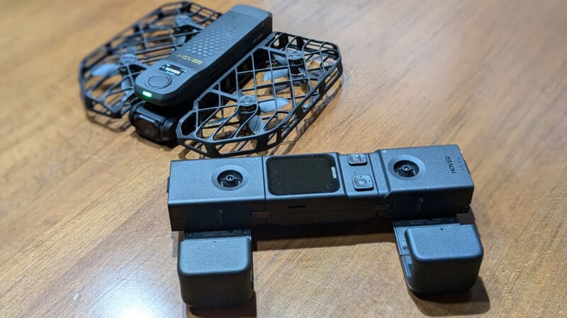 Two compact drones with protective cages are placed on a wooden surface. One drone is unfolded with its black frame visible. The other is folded, showcasing its sleek design and features. Both are marked with the brand "Hover.