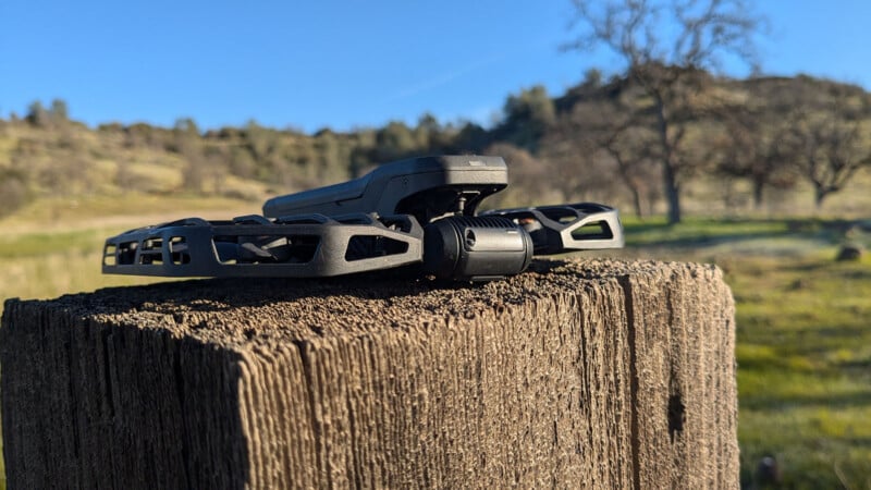 A drone rests on a weathered wooden post with a grassy landscape and trees in the background. The sky is clear and blue, suggesting a bright day. The focus is on the drone, highlighting its design and the texture of the wood.