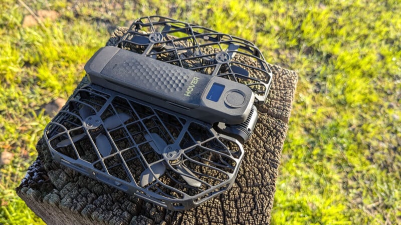 A black drone with protective mesh around its propellers sits on a wooden stump. The drone has a textured surface and the word "HOVER" is visible on it. The background shows green grass on a sunny day.