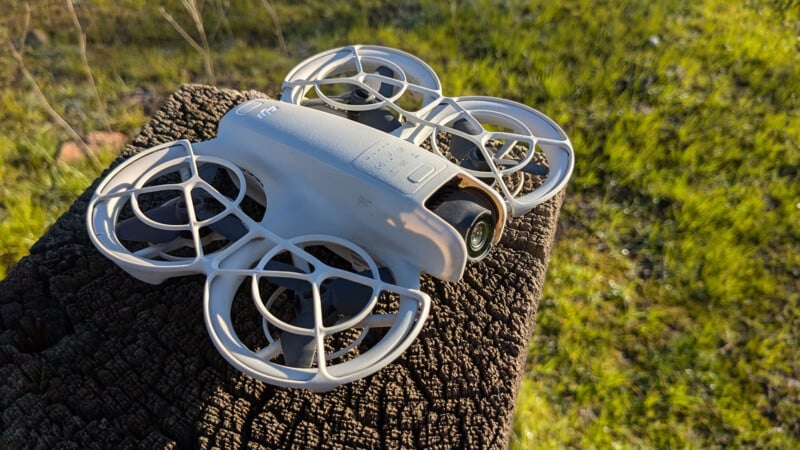 A white quadcopter drone with propeller guards rests on a weathered wooden surface. Sunlight highlights the drone against a grassy background, suggesting an outdoor setting.