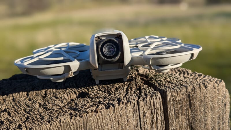 A small drone with a camera is perched on a weathered tree stump. The drone has a modern design with protective covers over its propellers. The background is a blurred, green field under bright sunlight.