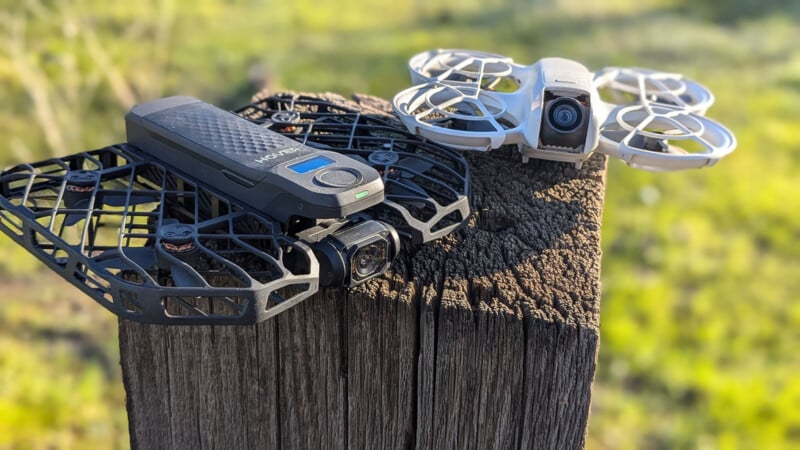 Two drones resting on a wooden post outdoors. One is black with a rectangular design, while the other is white with circular propeller guards. The background is grassy and blurred, suggesting a natural setting.