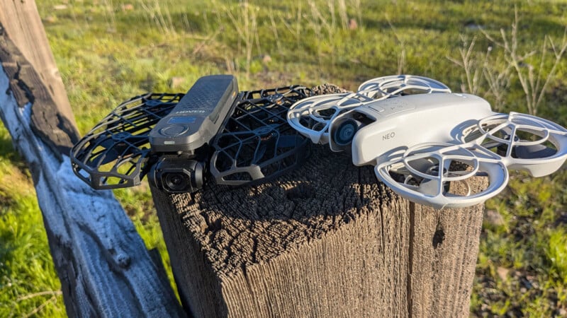 Two compact drones sit on a wooden post in a grassy area. The drone on the left is black with protective guards, and the one on the right is white with circular guards. Both are ready for flight.