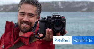 A person wearing a red jacket holds a Lumix camera towards the viewer, with a blurred body of water and snowy landscape in the background. The text "PetaPixel Hands-On" is displayed on the right side of the image.