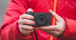 A person in a red jacket holds a black compact camera with both hands, pointing it towards the viewer. The camera lens is prominently visible, and the jacket features an orange zipper.