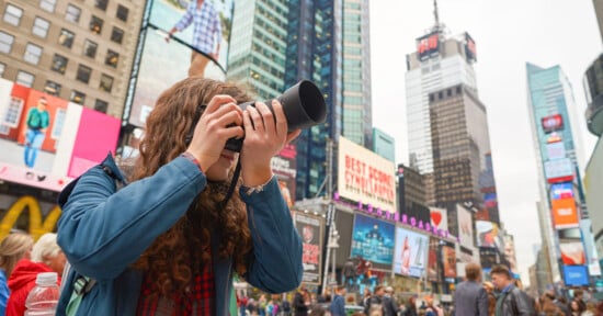 A person with long curly hair is taking a photo with a camera in a bustling urban area filled with tall buildings and large digital billboards. Crowds of people are visible, and the sky is overcast.