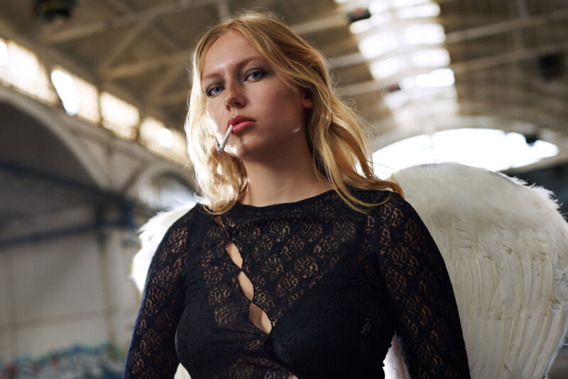 A person with long blonde hair wearing a black lace top stands in a large, industrial indoor setting. They have white angel wings and the light creates a halo effect around them. The ceiling has large, arched windows.