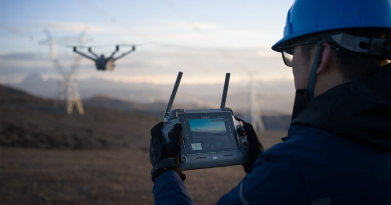 A person wearing a blue helmet and gloves operates a drone using a controller with a screen. The drone flies near power lines against a landscape of hills and a sunset sky.