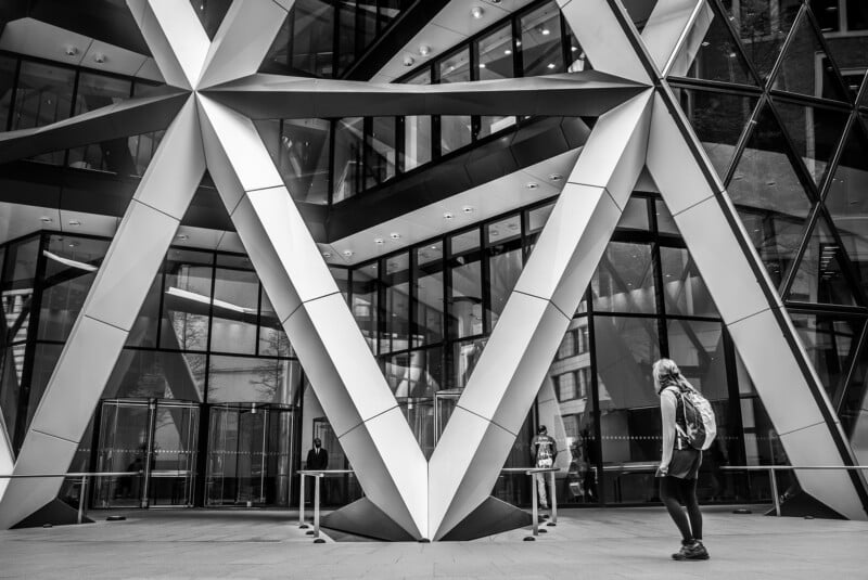A person with a backpack approaches a modern building with large, intersecting steel beams and glass walls. The architectural design features geometric patterns with triangular shapes. The scene is captured in black and white.
